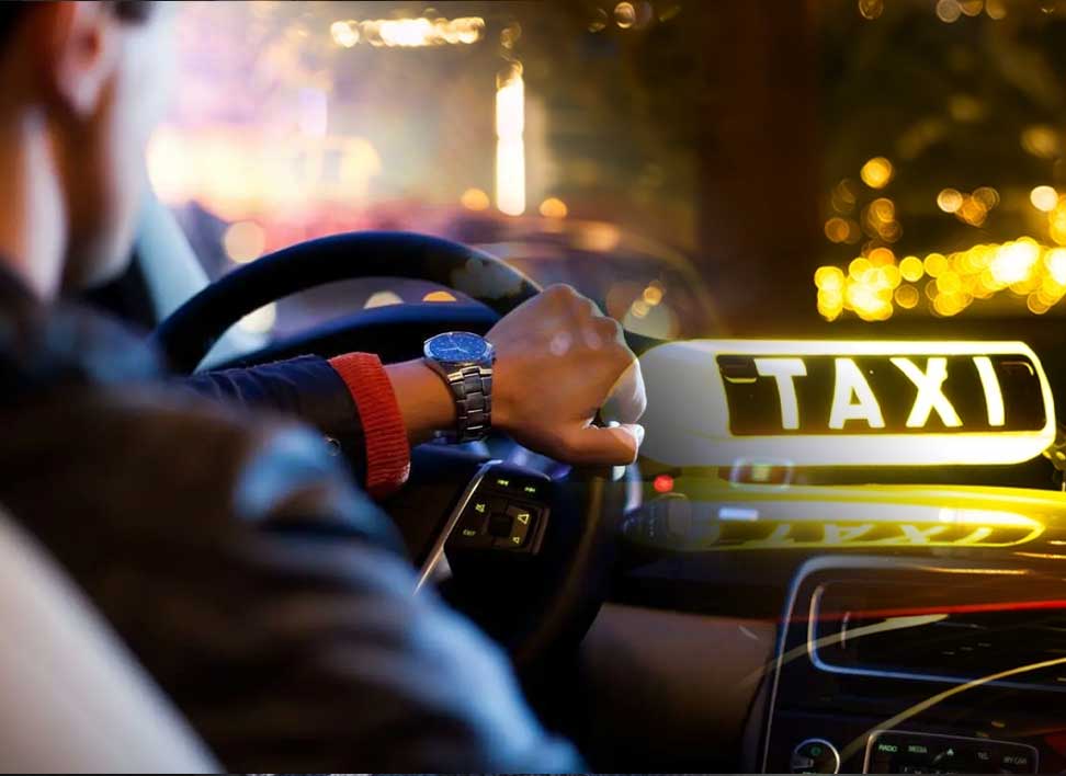 Conducteur au volant dans une voiture de taxi avec enseigne lumineuse "TAXI" à l’avant, ambiance nocturne.